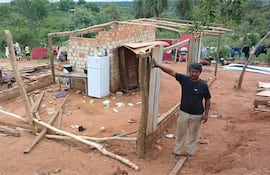 Hombre adulto con camiseta oscura y pantalones claros, en un sitio de construcción con materiales esparcidos y vegetación de fondo.