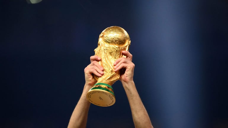 Copa del Mundo dorada sostenida en alto por manos en un ambiente de celebración, sin rostros visibles en el fondo.