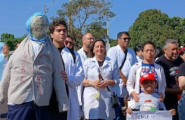 Grupo de médicos en batas blancas sostiene pancartas de protesta, un muñeco de 'Oscar Lovera' destaca en la escena.