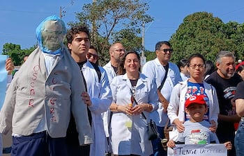 Grupo de médicos en batas blancas sostiene pancartas de protesta, un muñeco de 'Oscar Lovera' destaca en la escena.