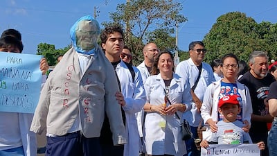 Grupo de médicos en batas blancas sostiene pancartas de protesta, un muñeco de 'Oscar Lovera' destaca en la escena.