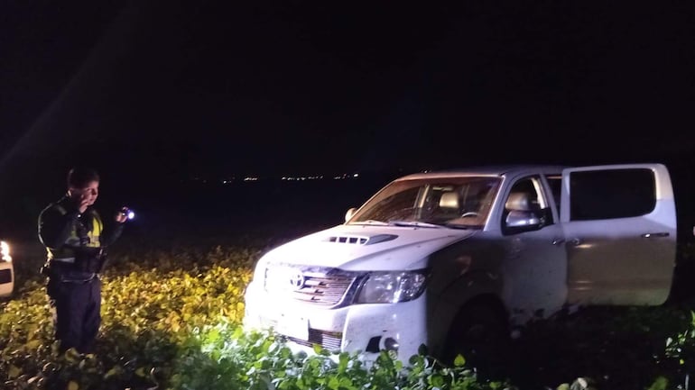 Policía con linterna junto a camioneta blanca con puerta abierta, en un ambiente oscuro y vegetación abundante.
