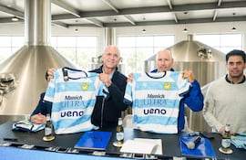 Hombres sosteniendo camisetas azules y blancas del Munich Rugby en sala con ambiente festivo y botellas de cerveza sobre la mesa.
