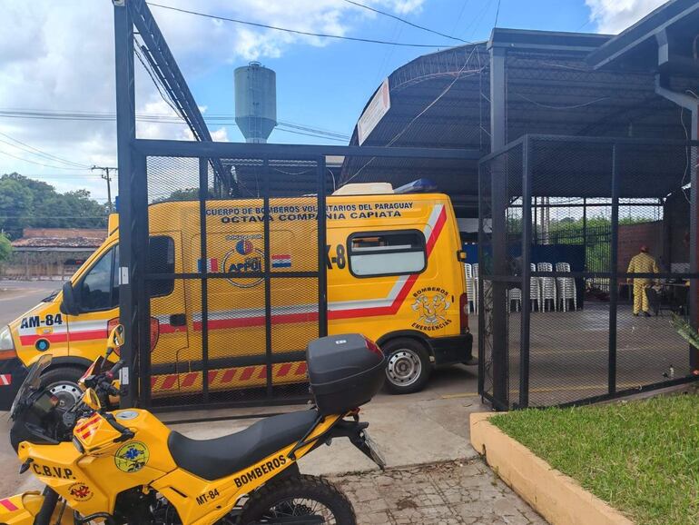 Ambiente exterior del cuartel de bomberos con moto amarilla y ambulancia, mostrando un bombero sentado en la parte trasera.