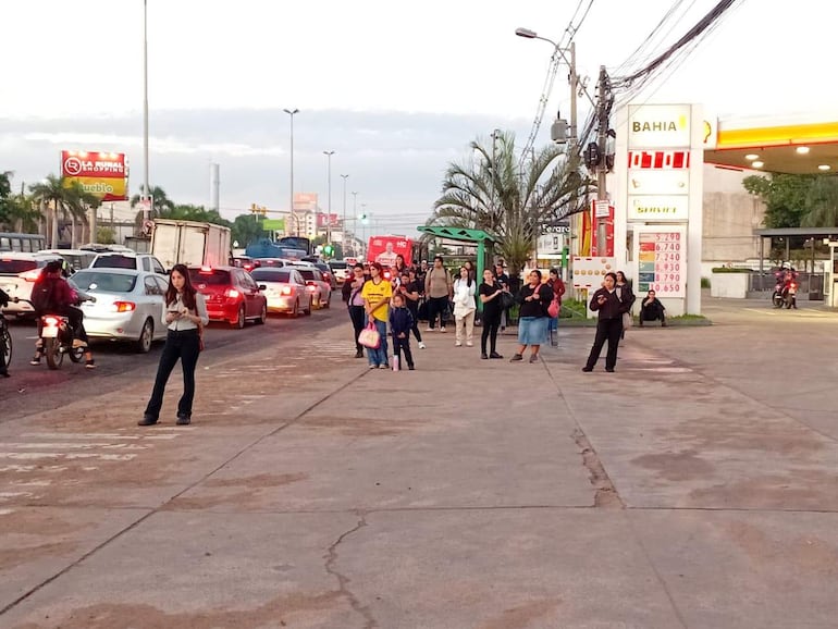 Grupo de personas esperando en la acera, algunos conversan, otros miran sus teléfonos, con tráfico intenso de fondo.