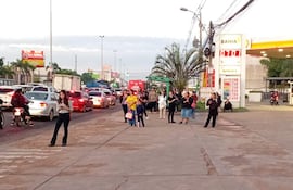 Grupo de personas esperando en la acera, algunos conversan, otros miran sus teléfonos, con tráfico intenso de fondo.