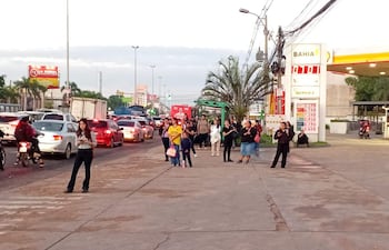 Grupo de personas esperando en la acera, algunos conversan, otros miran sus teléfonos, con tráfico intenso de fondo.