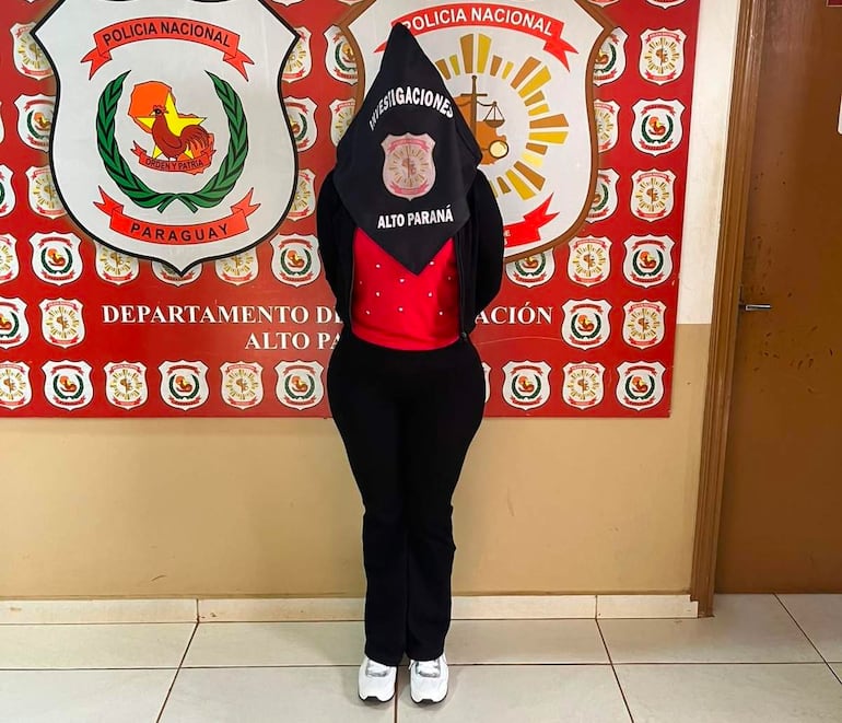 Persona vestida con pantalón negro y camiseta roja, con pañuelo negro que dice 'INVESTIGACIONES ALTO PARANÁ', en comisaría decorada con emblemas policiales.