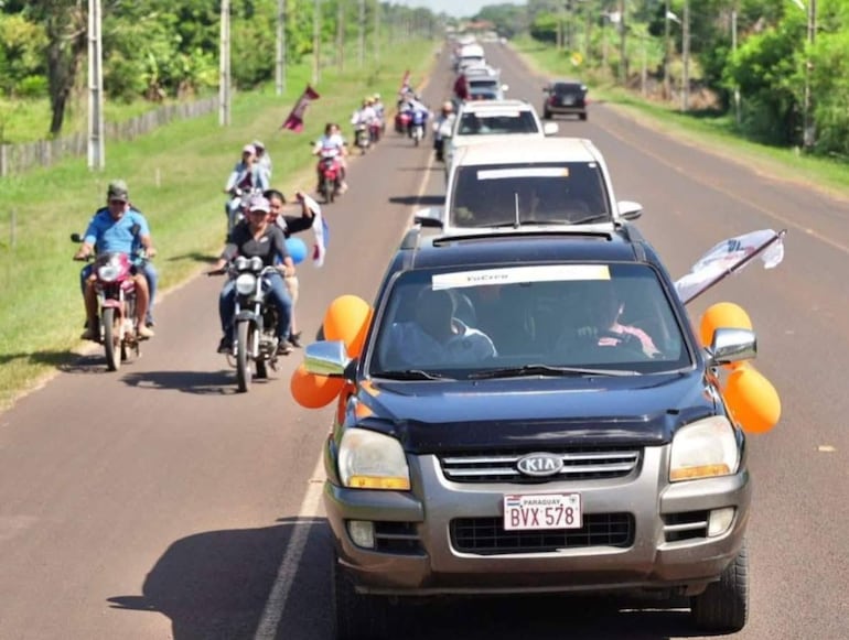 Caravana de vehículos y motocicletas decoradas con globos naranjas en una carretera rural, sin figuras públicas visibles.