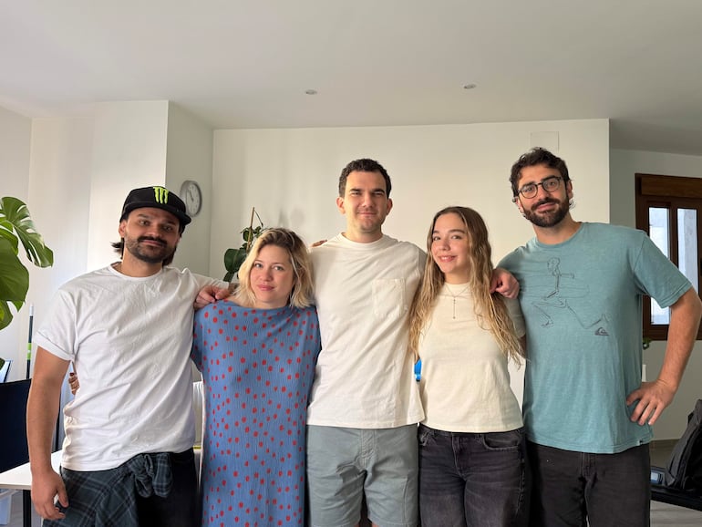 Grupo de cinco personas posando sonriendo; una mujer con vestido azul, hombres con camisetas y gorras en ambiente informal.