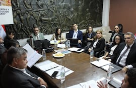 Andrea Picaso, vestida de forma ejecutiva, escucha atentamente a un hombre con gafas en una reunión con otros asistentes alrededor de una mesa.