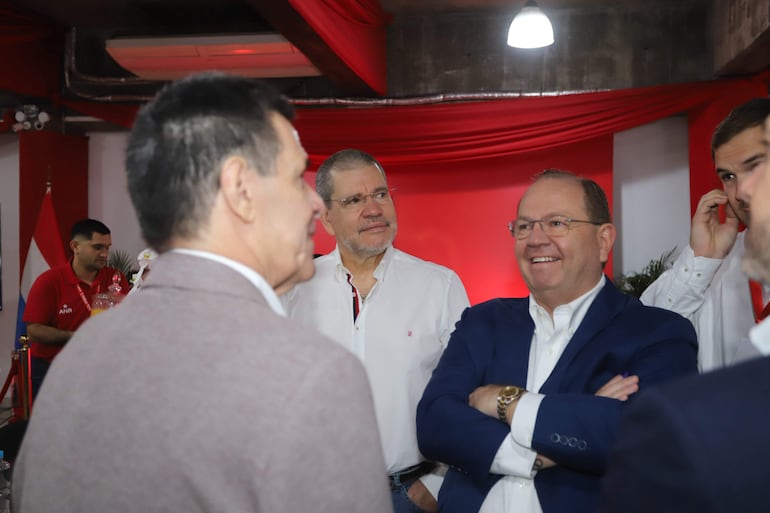 Horacio Cartes sonríe con chaqueta azul, junto a Camilo Pérez y Antonio Carlos Barrios, en un ambiente decorado con banderas paraguayas.