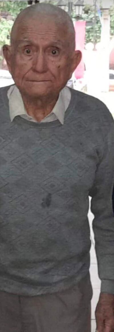 Hombre mayor de pie con suéter gris y camisa blanca, con expresión neutra en un ambiente interior iluminado.