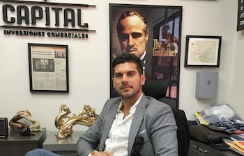 Hombre con traje gris claro sentado frente a escritorio, rodeado de documentos y figura dorada de dragón en oficina decorada.