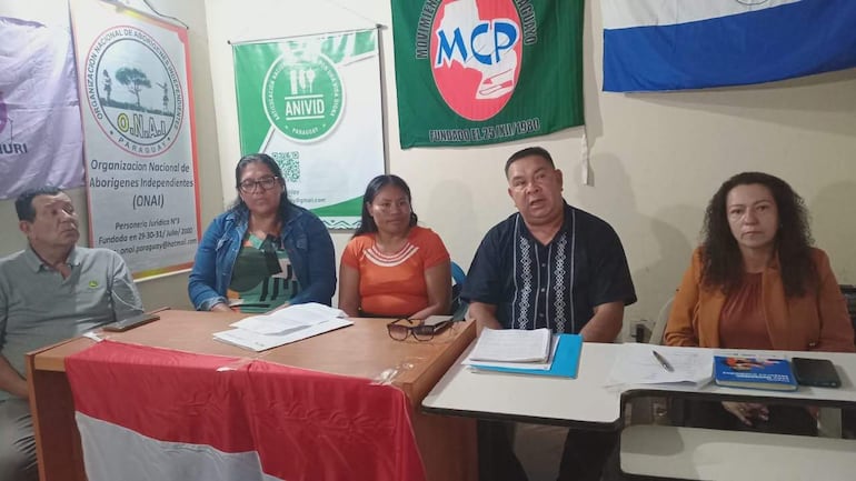 Hombre con chaqueta marrón habla en mesa, rodeado por cuatro personas, con banderas en el fondo y documentos sobre la mesa.