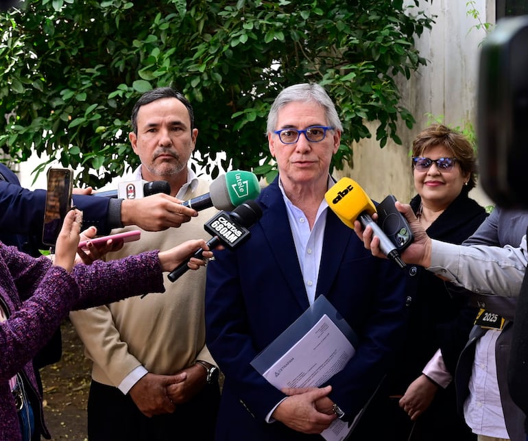 Emiliano Fernández con gafas y documentos, mientras César Ruiz Díaz sonríe, rodeados de micrófonos en un ambiente verde.