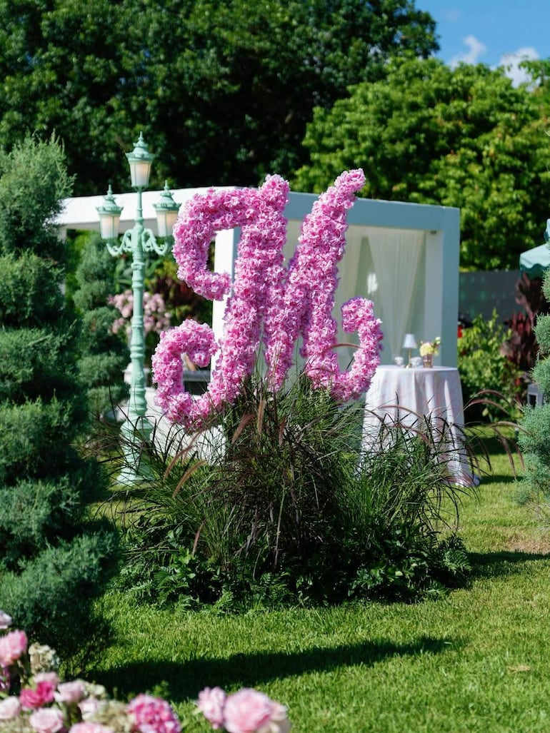 Gran arreglo floral en forma de 'M' con flores rosas en un jardín adornado, rodeado de vegetación y mesas preparadas.
