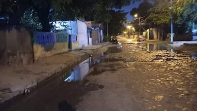 Calle empedrada con charcos reflejando luz de farolas, y un automóvil estacionado al fondo. No hay personas en la escena.