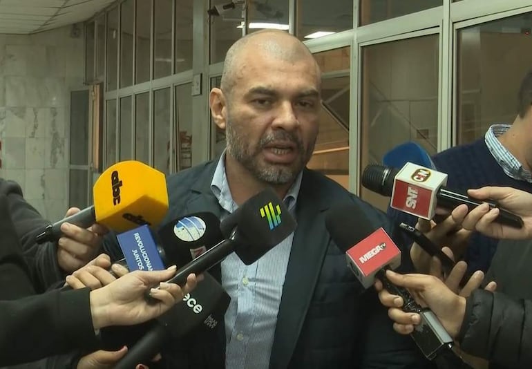 Hugo Ramírez, hombre de mediana edad con chaqueta oscura y barba, responde preguntas rodeado de micrófonos de prensa en un entorno con paredes de mármol.