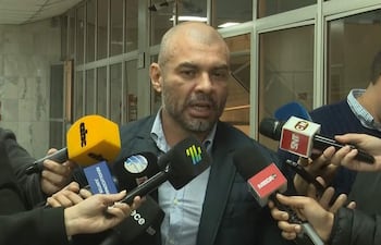 Hugo Ramírez, hombre de mediana edad con chaqueta oscura y barba, responde preguntas rodeado de micrófonos de prensa en un entorno con paredes de mármol.