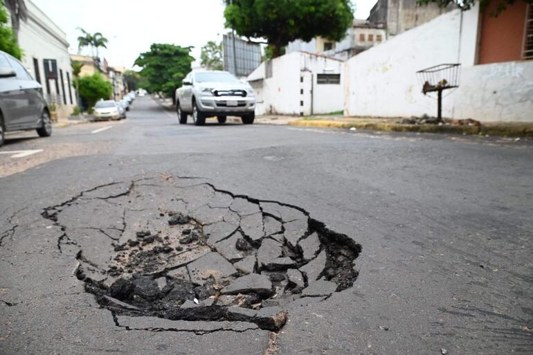 Calle urbana con gran bache en primer plano, vehículos estacionados a un lado y árboles al fondo.