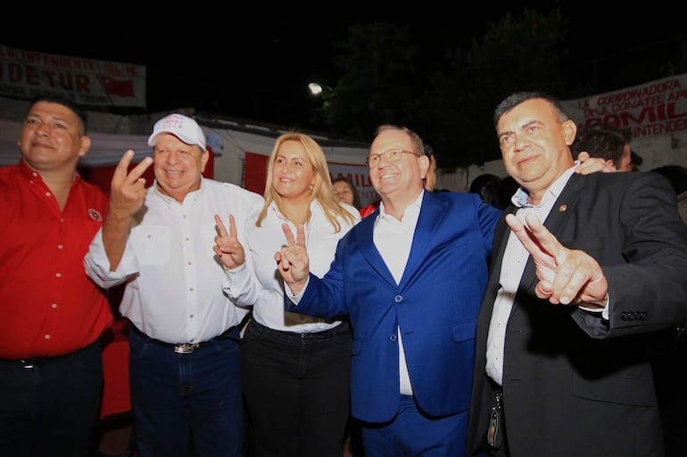 Camilo Pérez y cinco personas sonríen, posando en un evento con ropa formal en tonos blancos y rojos, levantando dos dedos.