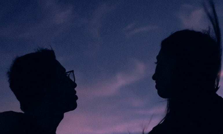 Silhuetas de un hombre con gafas y una mujer de cabello largo conversan al atardecer en un cielo púrpura y azul.