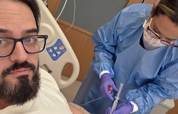 Hombre con barba y gafas en camiseta blanca toma una selfie mientras recibe atención médica de una mujer con mascarilla y uniforme azul.