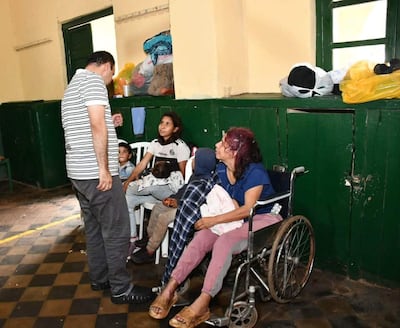 Hombre de pie conversa con mujer sentada en un albergue, mientras un niño escucha atento. Ambiente desordenado con sillas y bolsas.