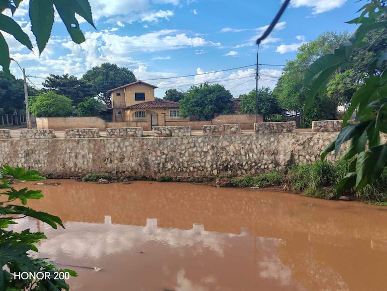Paisaje urbano con río marrón, casa amarilla y árboles, en un ambiente residencial tranquilo.