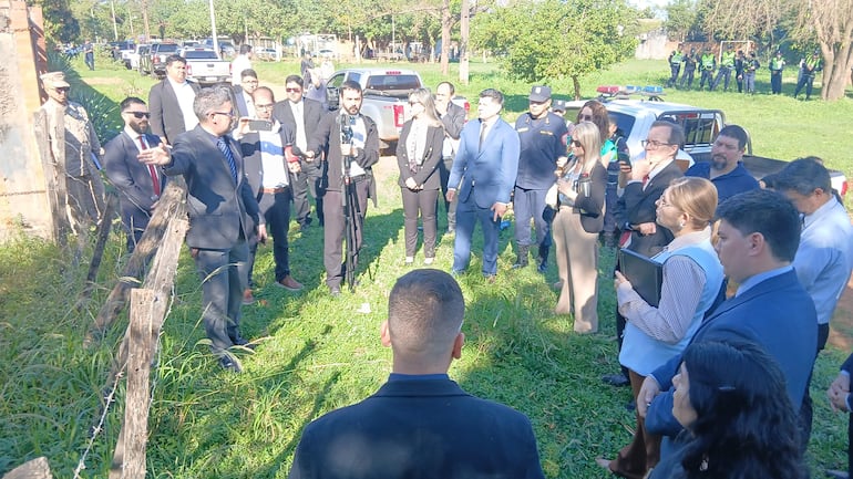 Fermín Segovia en traje negro gesticula frente a un grupo en un área verde, rodeado de autos estacionados y miembros de la policía.
