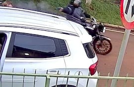 Automóvil blanco en primer plano, motocicleta negra detrás con conductor que lleva casco, ambiente urbano con vegetación y señal de tránsito.