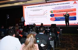 Hombre presentando en escenario con gráfico sobre precios de combustibles, audiencia atenta en un salón del hotel.
