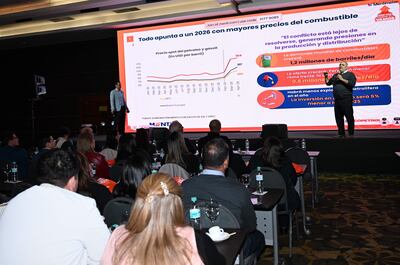 Hombre presentando en escenario con gráfico sobre precios de combustibles, audiencia atenta en un salón del hotel.