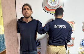 Alfredo Ortigoza con camiseta oscura, de pie con expresión seria, mientras un agente de Interpol lo acompaña de espaldas.