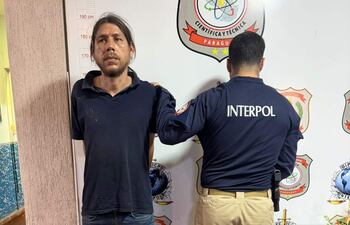 Alfredo Ortigoza con camiseta oscura, de pie con expresión seria, mientras un agente de Interpol lo acompaña de espaldas.