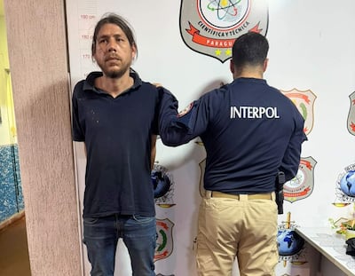 Alfredo Ortigoza con camiseta oscura, de pie con expresión seria, mientras un agente de Interpol lo acompaña de espaldas.
