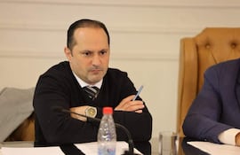 Hombre delgado con suéter negro y corbata, sentado en mesa con documentos, en ambiente formal de reunión.