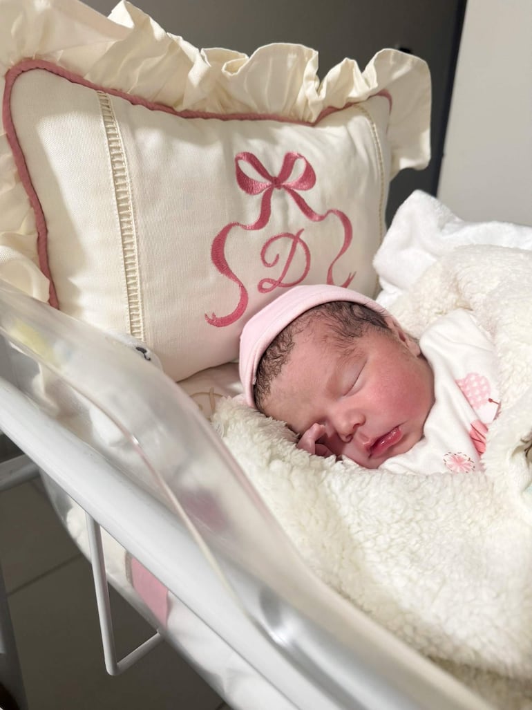 Bebé dormida en cuna blanca, cubierta con manta suave y gorro rosa, en habitación con paredes claras.