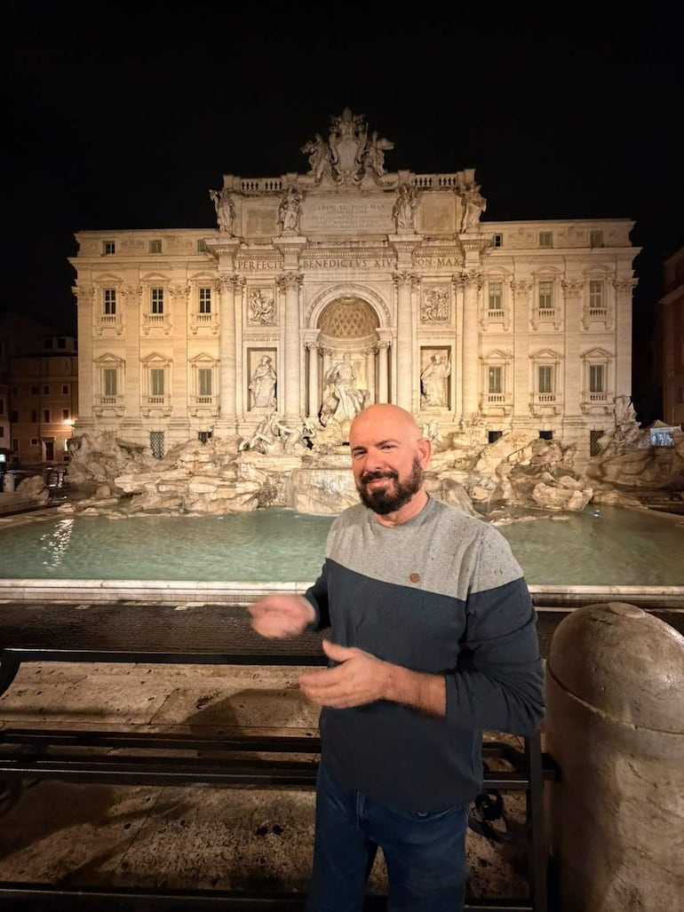 Hombre con barba, camiseta gris oscuro y azul, gesticulando ante la iluminada Fontana di Trevi en un ambiente nocturno.