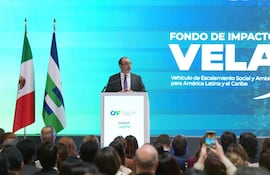 Hombre en traje oscuro y gafas habla tras un podio, con banderas de México y un gran panel detrás con el evento.