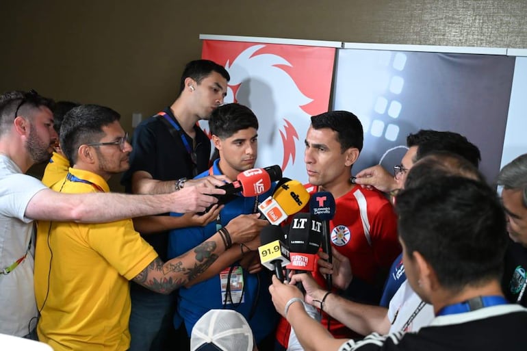 Junior Alonso en camiseta roja con detalles en blanco, respondía preguntas rodeado de periodistas con micrófonos en un ambiente de conferencia de prensa.