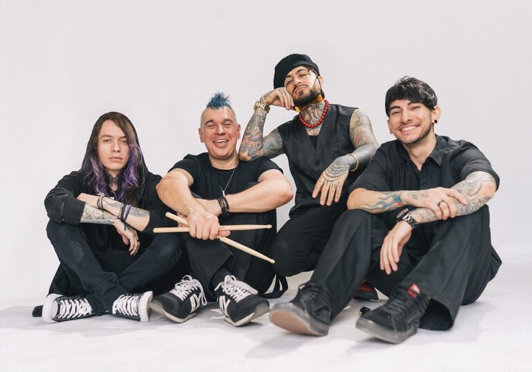 Cuatro hombres sonrientes, con estilos oscuros y tatuajes, posan relajados sobre fondo blanco en un estudio.