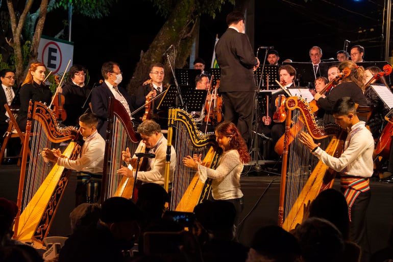 Cuatro arpistas, vestidos con trajes tradicionales, tocan sus instrumentos mientras un público concentrado escucha atentamente en un ambiente nocturno.