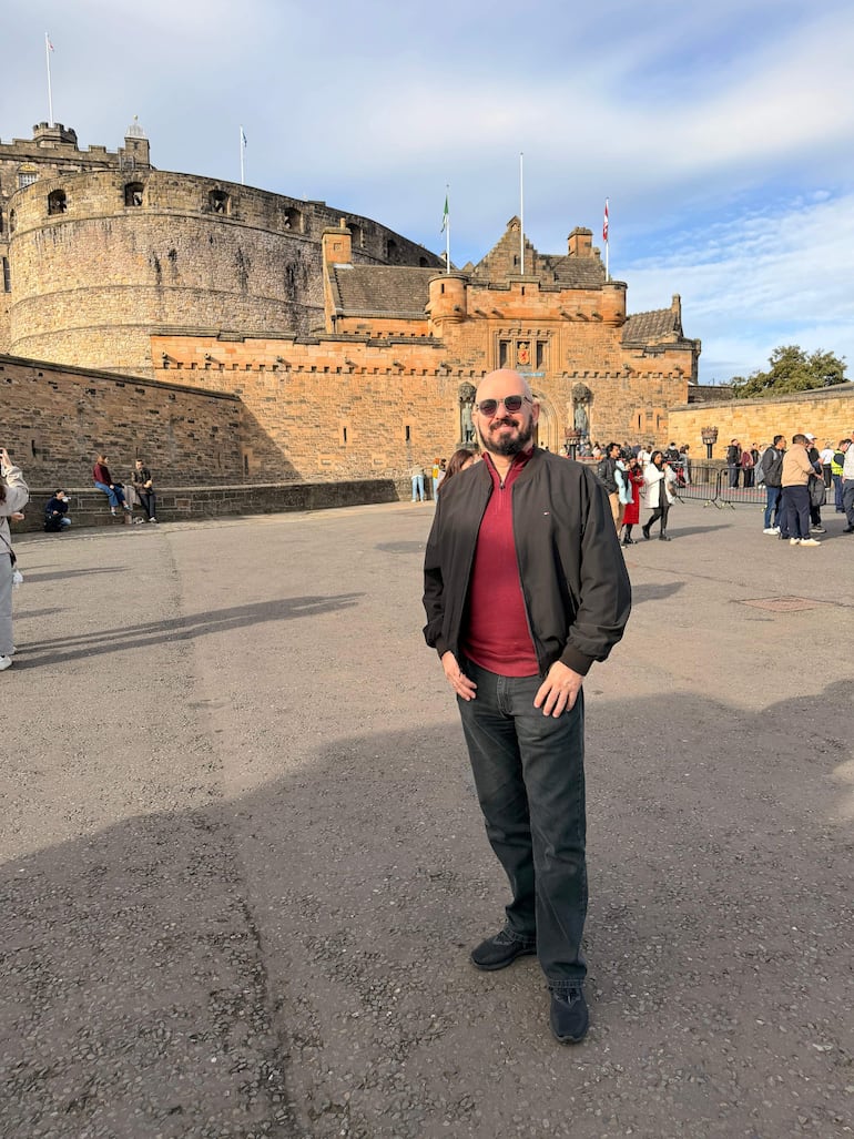 Hombre con barba y gafas oscuras, vestido con camiseta roja y chaqueta negra, frente al Castillo de Edimburgo.