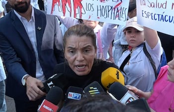 Rosanna González con camiseta negra, gesticulando intensamente rodeada de micrófonos, mientras personas sostienen carteles en protesta.