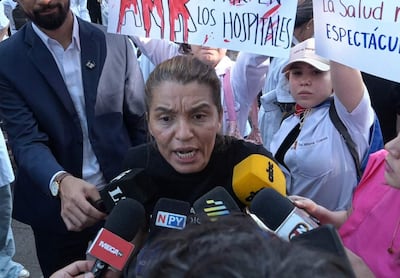 Rossana González con camiseta negra, gesticulando intensamente rodeada de micrófonos, mientras personas sostienen carteles en protesta.