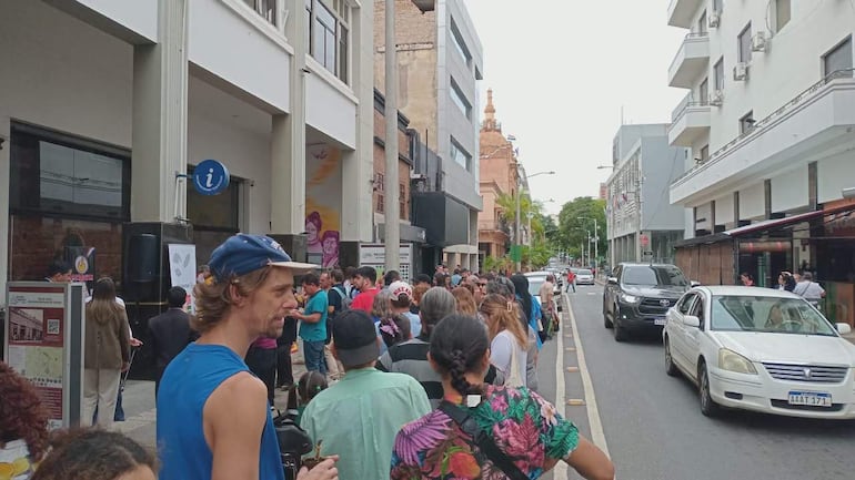 Multitud alineada en la vereda, esperando por degustación, con variedad de camisetas y edificios de fondo.