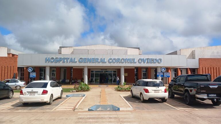 Fachada del Hospital General de Coronel Oviedo con vehículos estacionados, en un día soleado y con nubes.