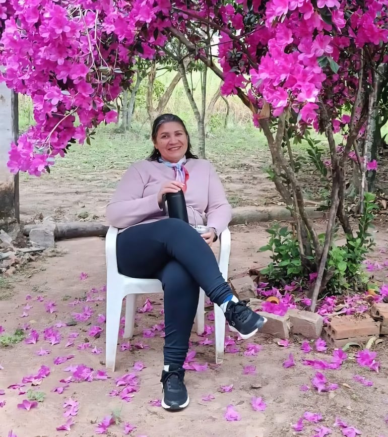 Albina Cardozo sonriente, vestida con suéter claro y leggings oscuros, sentada en una silla plástica blanca rodeada de flores rosadas.